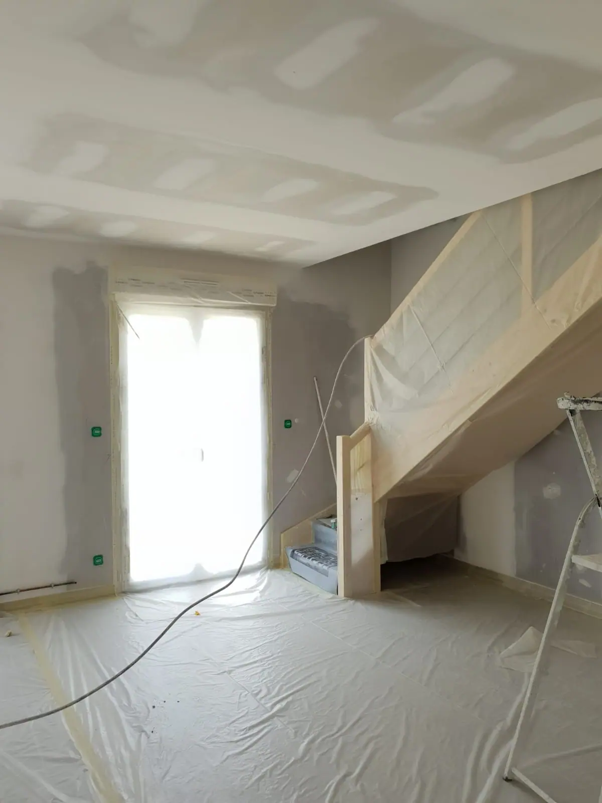 Escalier en bois emballé sous film de protection, porte-fenêtre masquée et sol bâché dans une pièce aux plaques de plâtre jointées