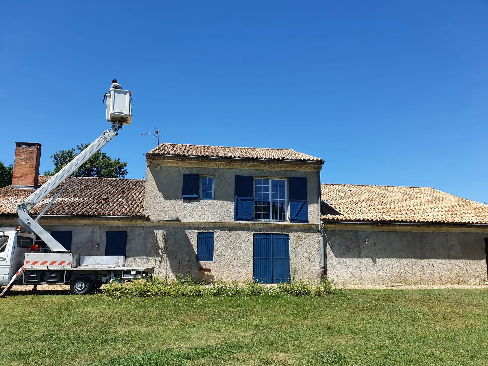 Intervention en hauteur : nacelle élévatrice déployée devant une maison de campagne aux volets bleus et aux toits de tuiles