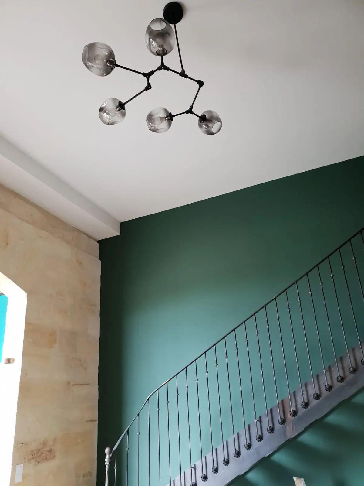 Montée d’escalier à rampe métallique le long d’un mur vert profond, plafond blanc, suspension à globes de verre et mur en pierre de taille
