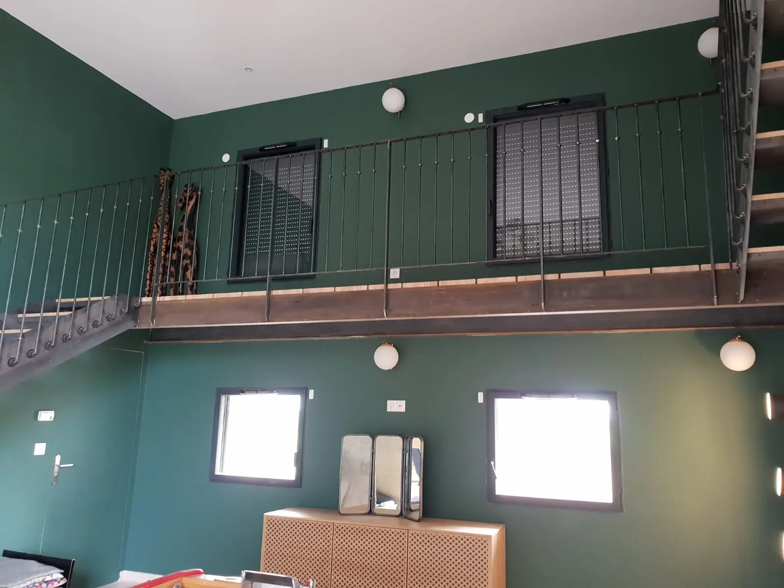 Après : séjour en double hauteur aux murs vert profond et au plafond blanc, sous une mezzanine à garde-corps en métal