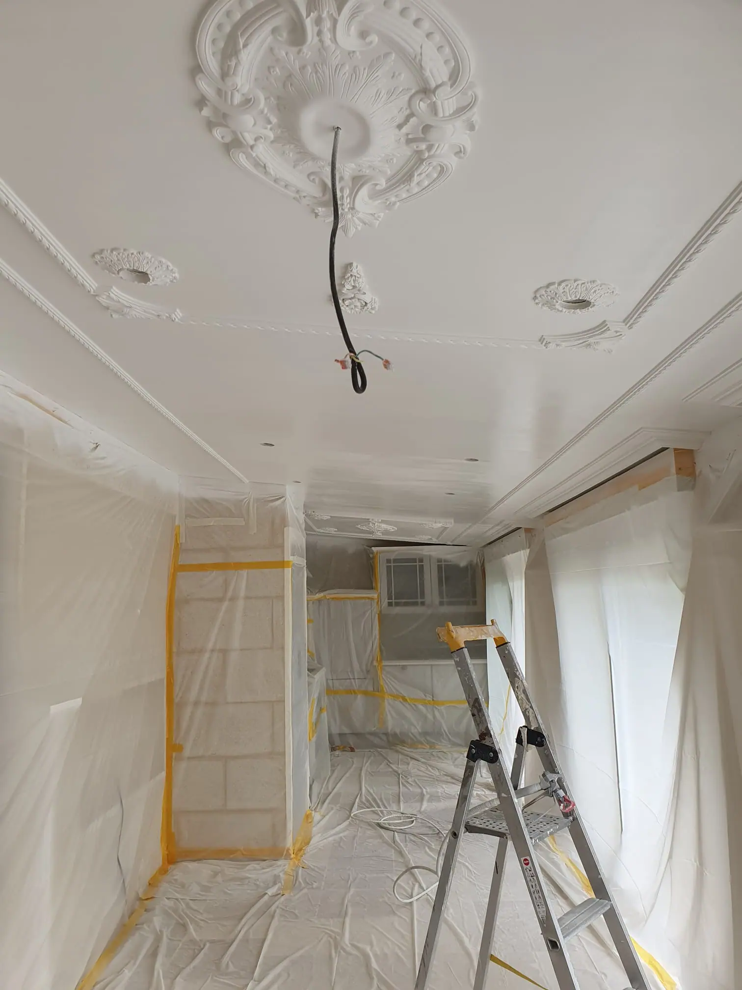 Le même plafond vu depuis l’autre extrémité de la pièce : rosaces et corniches en plâtre au-dessus de murs sous bâches