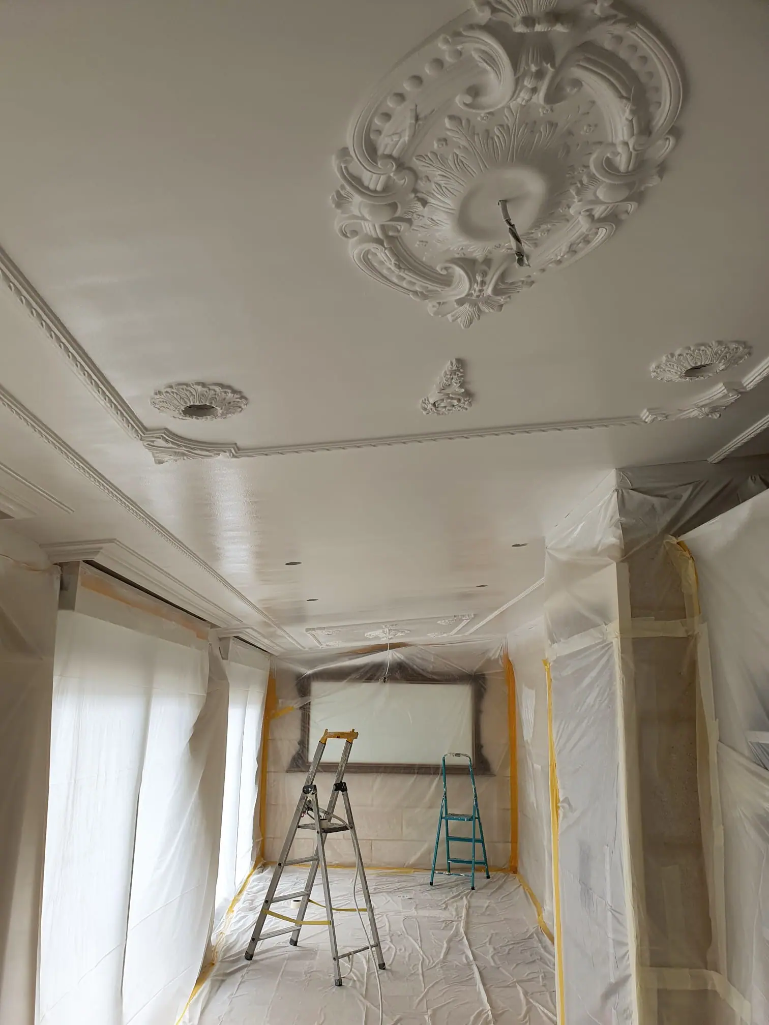 Pièce entièrement protégée avant peinture : murs, fenêtres et sol sous bâches et ruban de masquage, sous un plafond à rosaces
