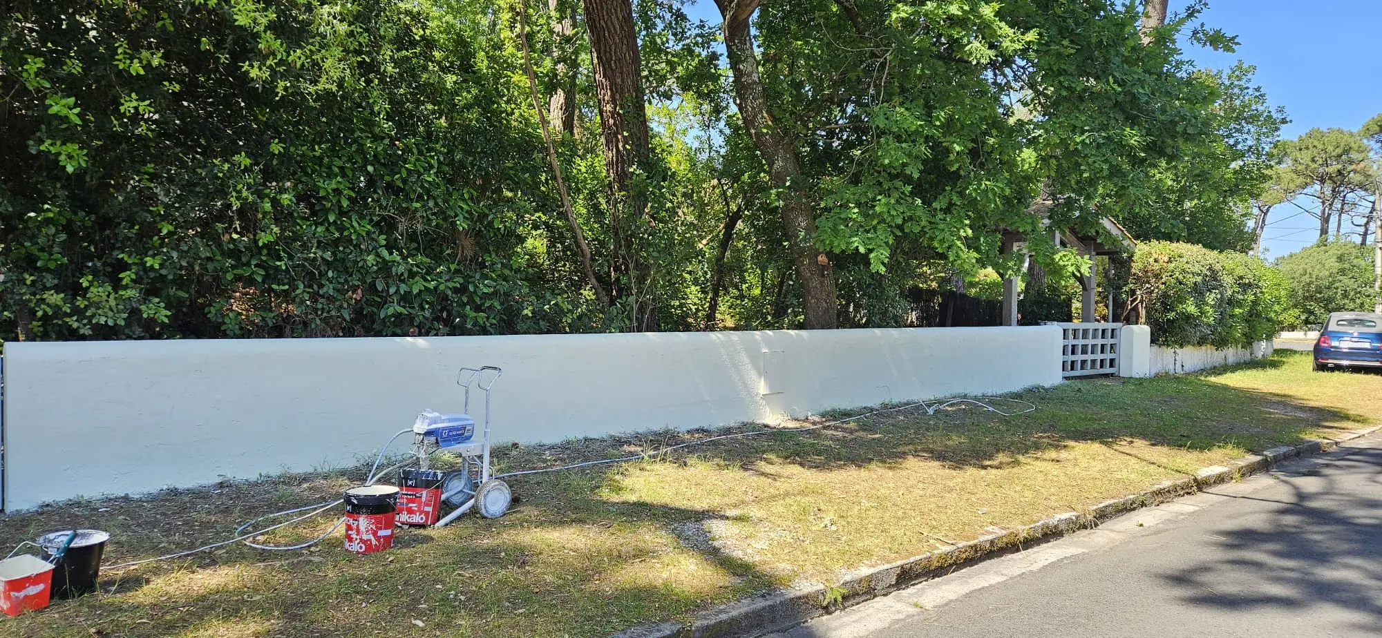 Le mur de clôture repeint en blanc jusqu’au portillon, pompe airless et seaux de peinture sur l’accotement