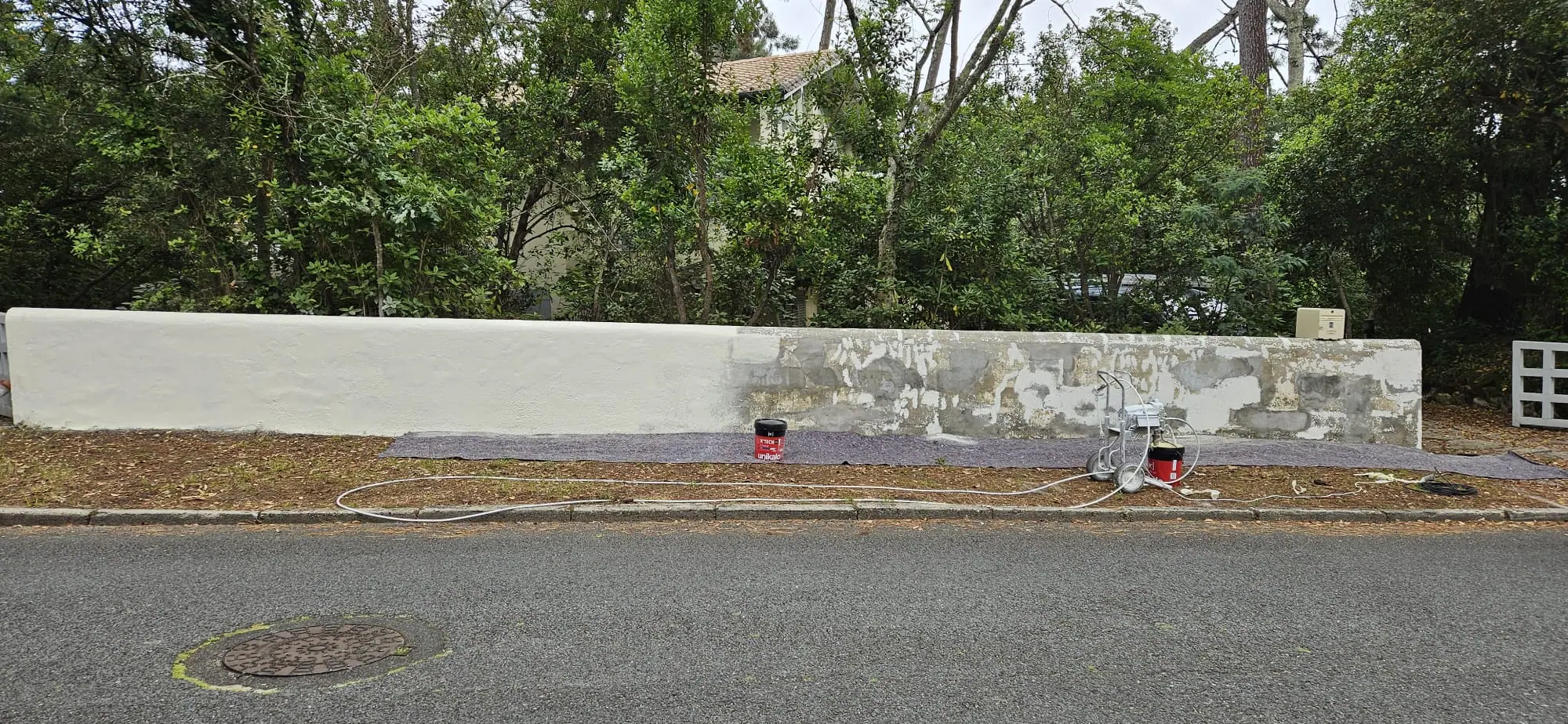 Mur de clôture en cours de mise en peinture : partie déjà repeinte en blanc, reprises d’enduit sur l’ancienne peinture écaillée, feutre de protection et pompe airless au pied du mur
